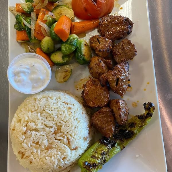Mediterranean dish with grilled meat, rice, roasted vegetables, a grilled pepper, and a side of sauce.