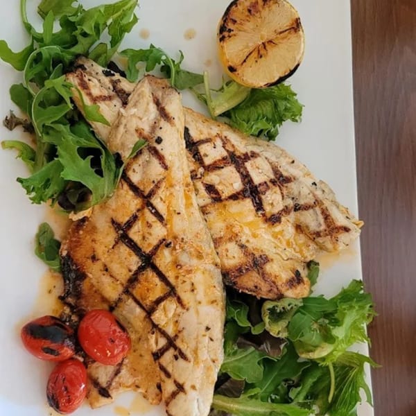 Grilled fish fillets with char marks, served with fresh greens, roasted tomatoes, and a lemon half.